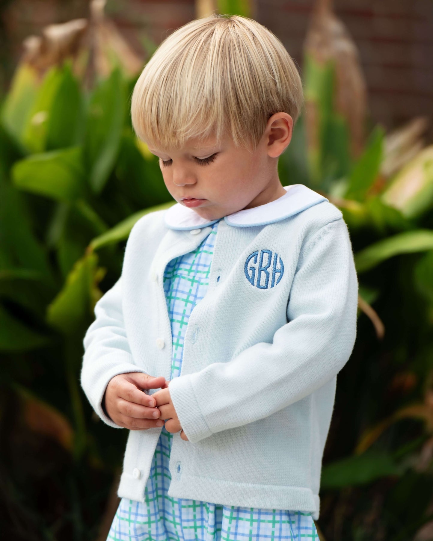 Young boy wearing a light blue jacket with monogrammed initials, standing outdoors.