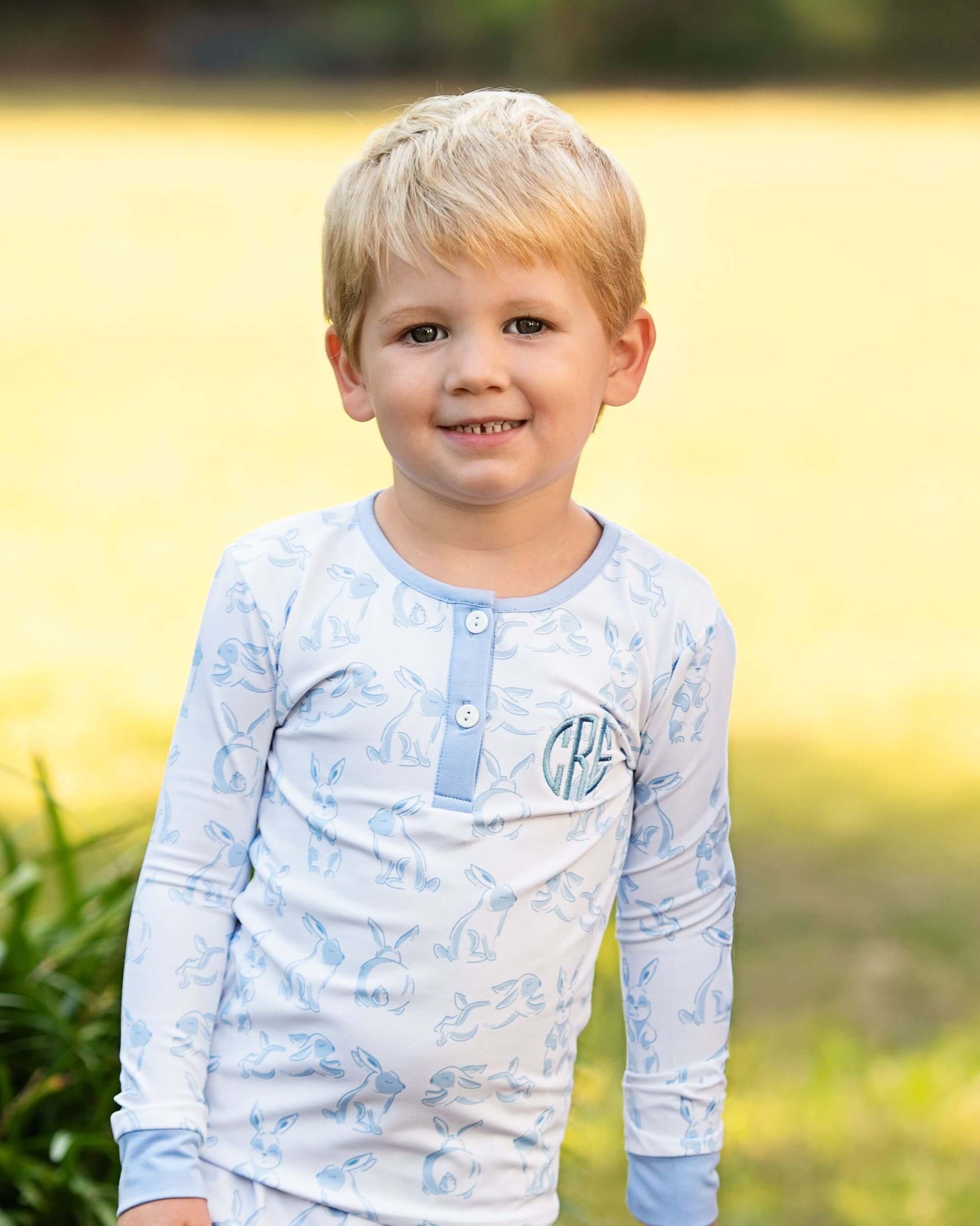 Young boy wearing a light blue long-sleeve shirt with a pattern, standing outdoors with a blurred natural background.