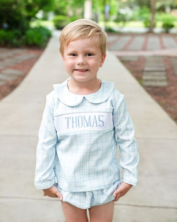 Young boy wearing a light blue outfit with 'THOMAS' printed on it, standing outdoors.