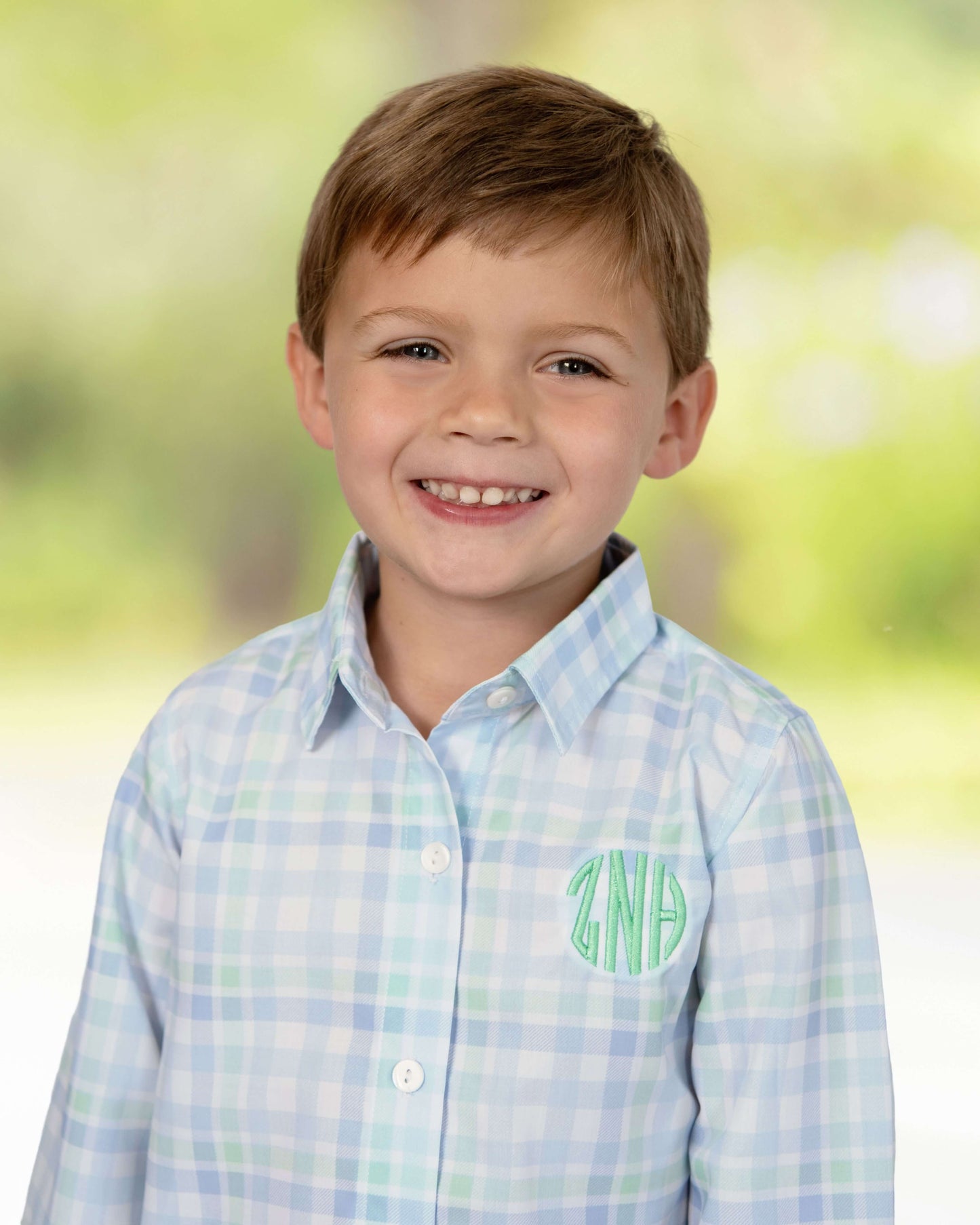 Young boy wearing a light blue plaid shirt with a monogram against a blurred green background