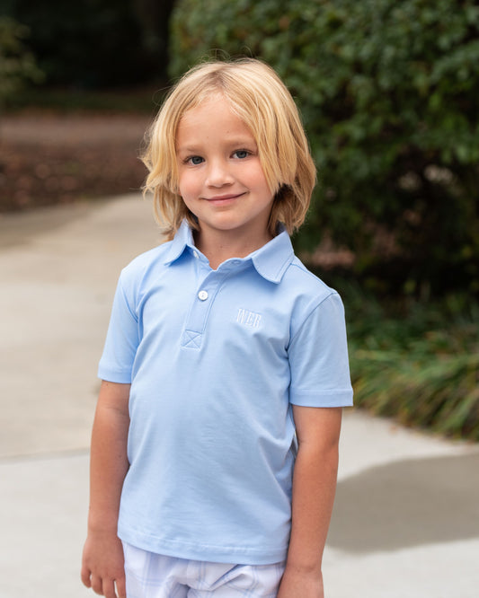 Young boy wearing a light blue polo shirt standing outdoors with greenery in the background