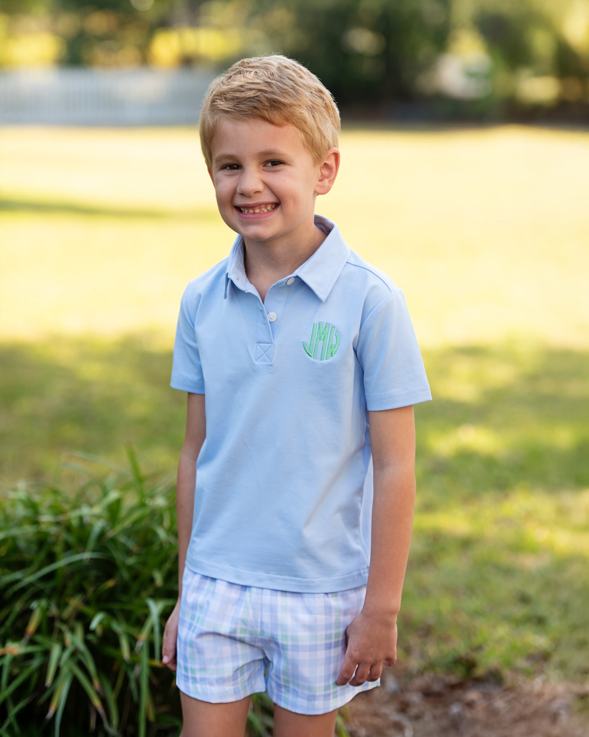 Young boy wearing a light blue polo shirt with a monogram and checkered shorts standing outdoors.