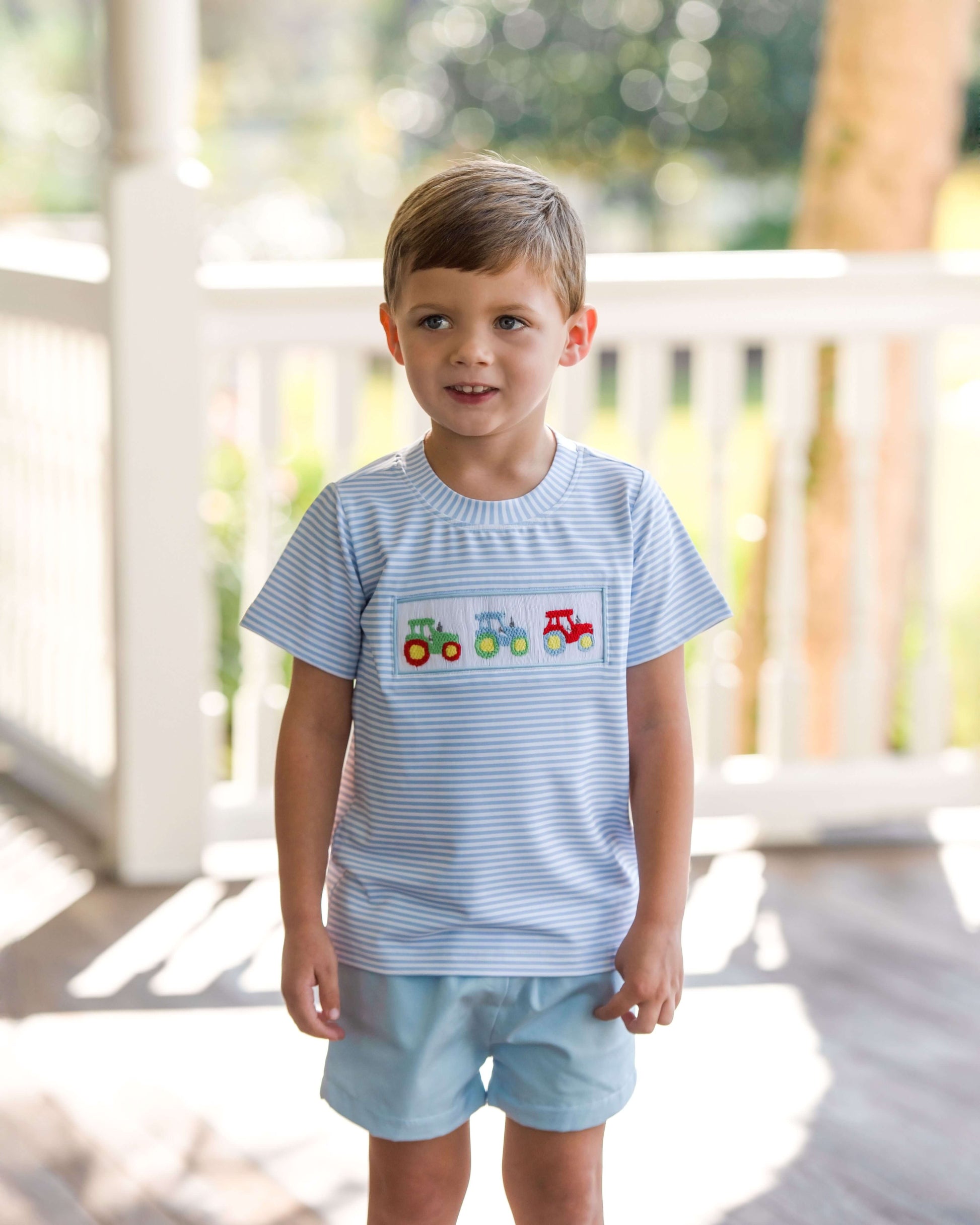 Young boy wearing a light blue striped shirt with train design and light blue shorts on a wooden deck.