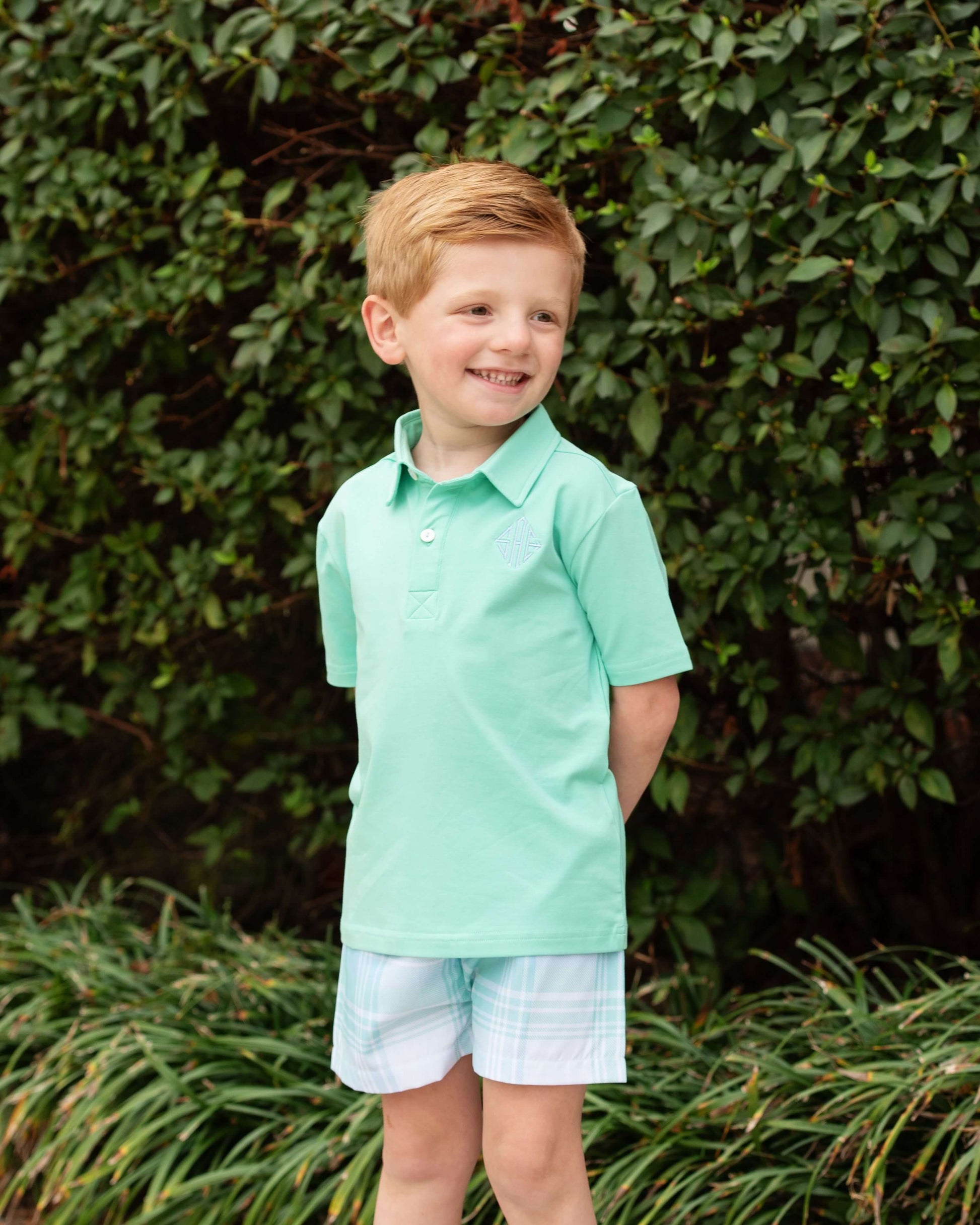 Young boy wearing a light green polo shirt and plaid shorts standing in front of green foliage.