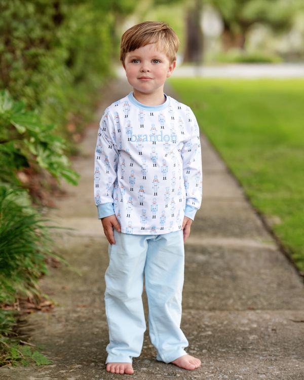 Young boy wearing a patterned long-sleeve shirt and light blue pants standing on a path with greenery.