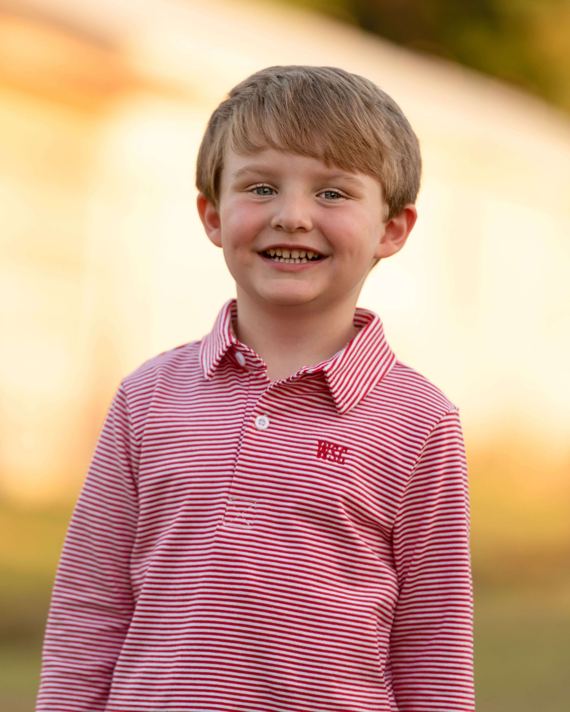 Young boy wearing a red and white striped shirt with a logo, standing outdoors with a blurred background.