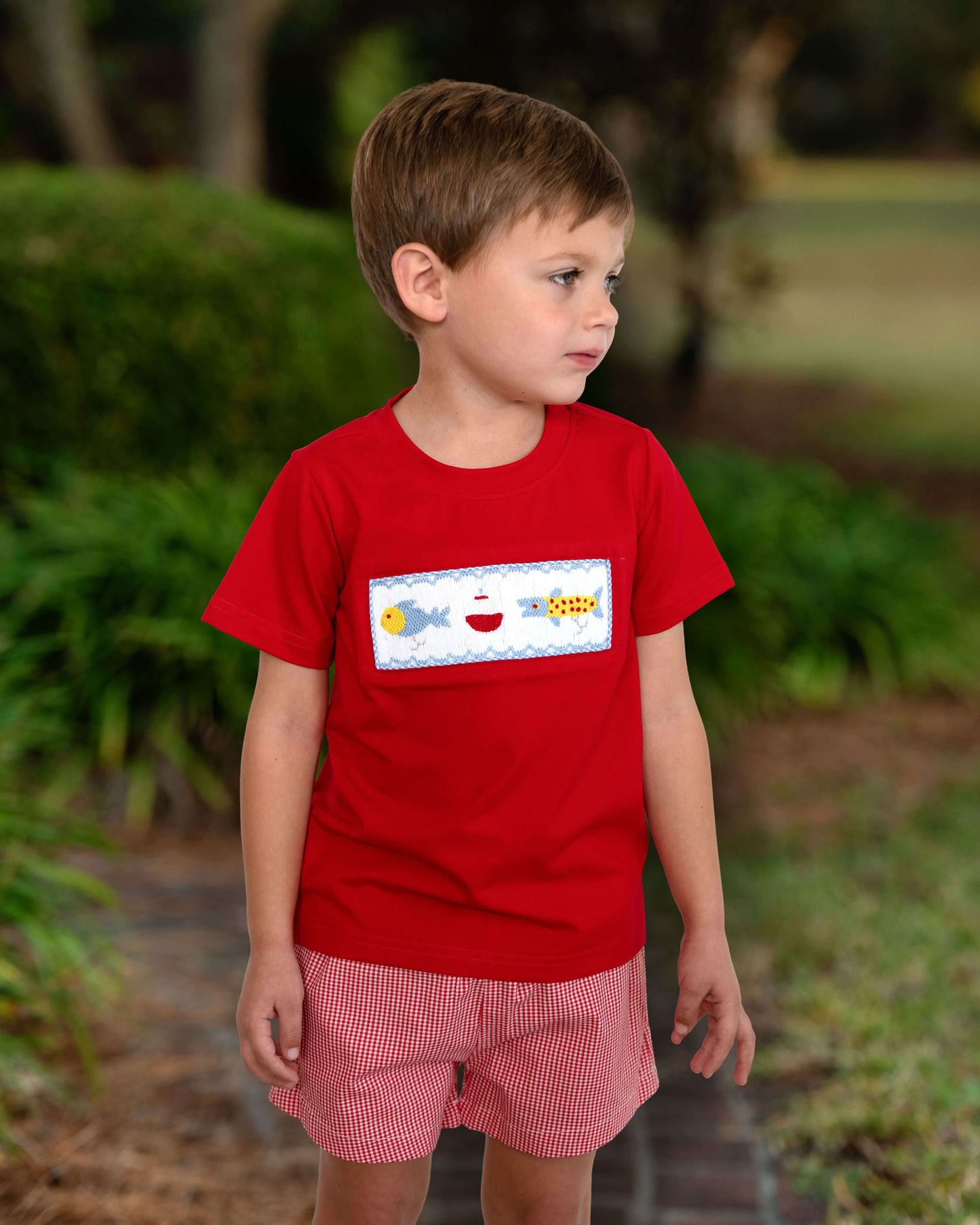 Young boy wearing a red shirt with a colorful graphic and pink shorts outdoors.