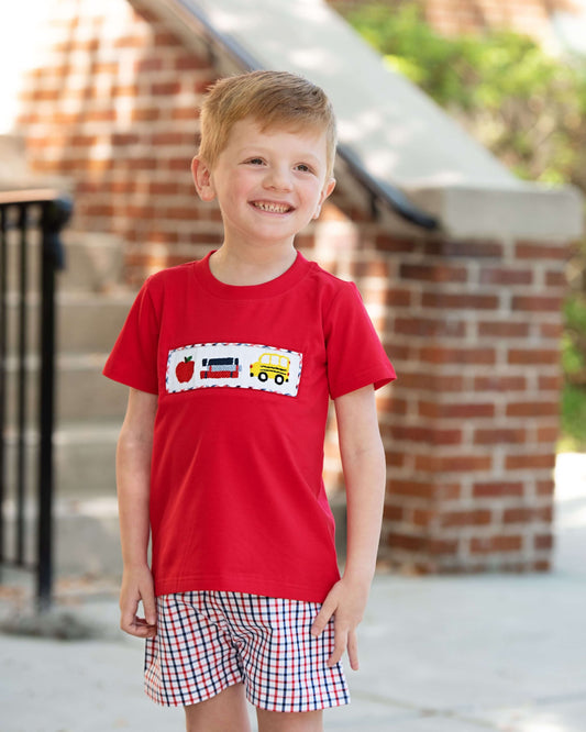 Young boy wearing a red shirt with a design and plaid shorts standing outdoors.