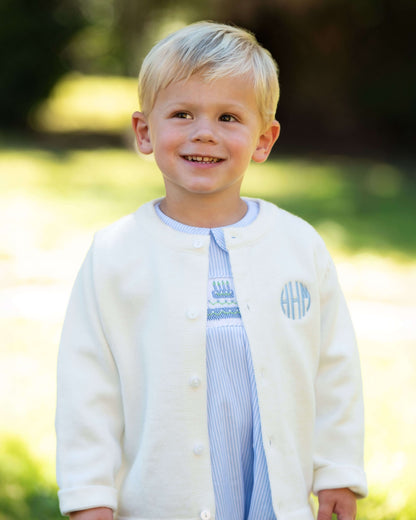 Young boy wearing a white cardigan with a monogram, standing outdoors.