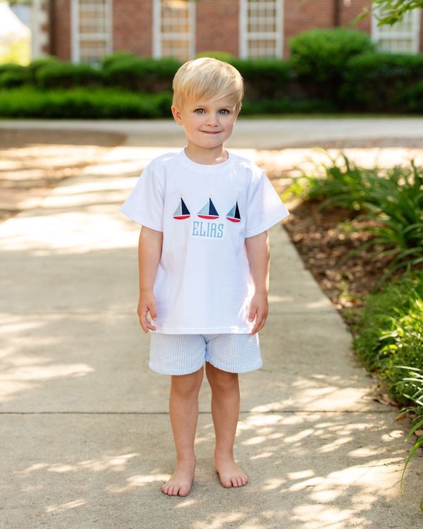 Young boy wearing a white t-shirt with sailboat design and shorts, standing on a sidewalk.