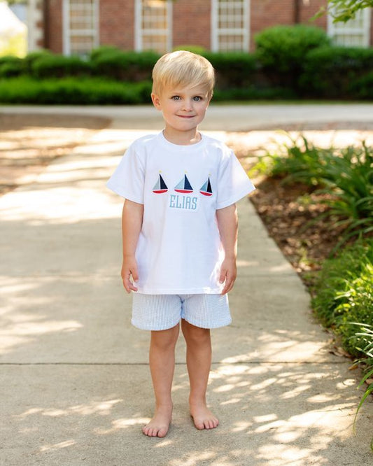 Young boy wearing a white t-shirt with sailboat design and shorts, standing on a sidewalk.