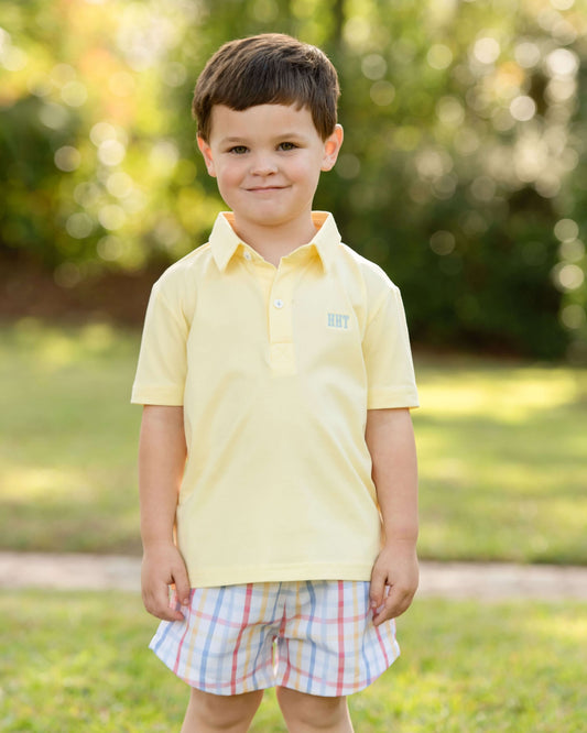 Young boy wearing a yellow polo shirt and plaid shorts standing outdoors.