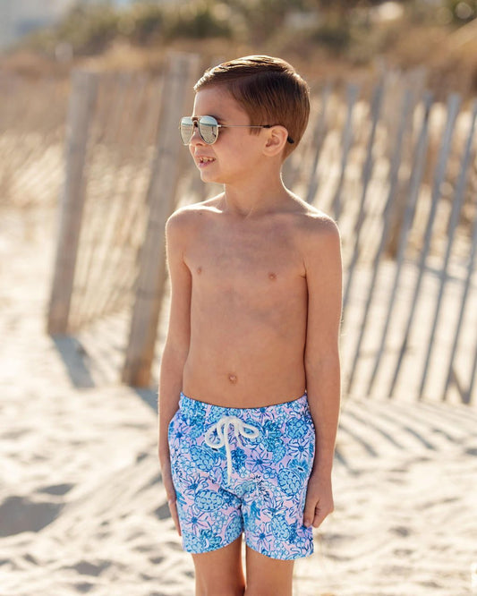 Young boy wearing blue floral swim shorts on a sandy beach.