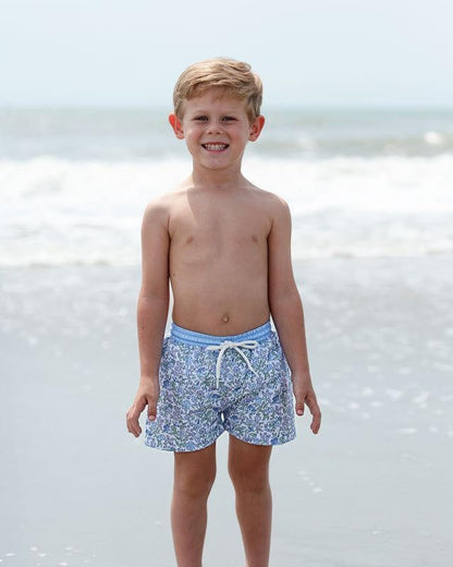 Young boy wearing blue floral swim shorts standing on a beach.
