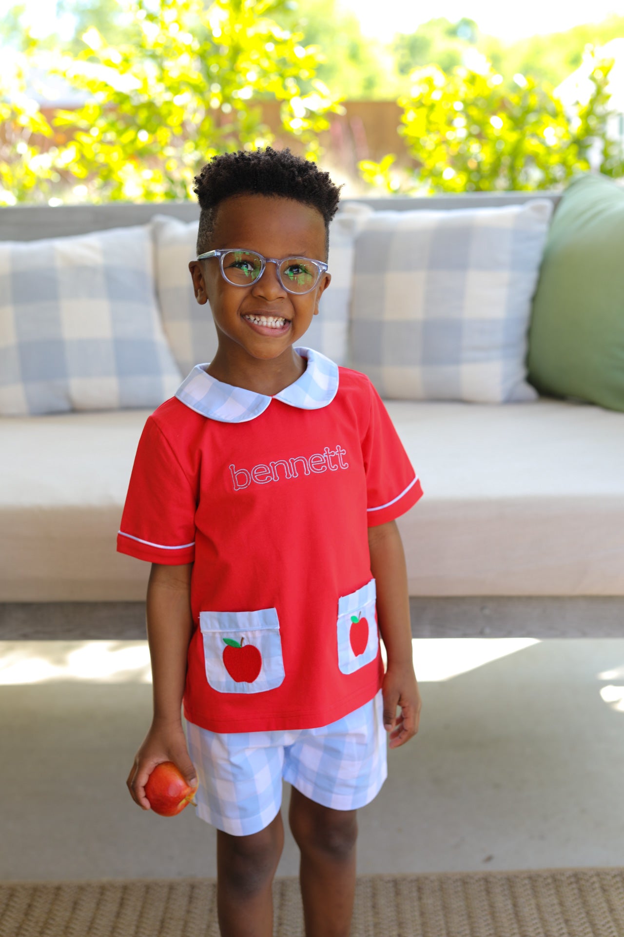 Young boy wearing blue gingham shorts and red top with apples on the pockets.