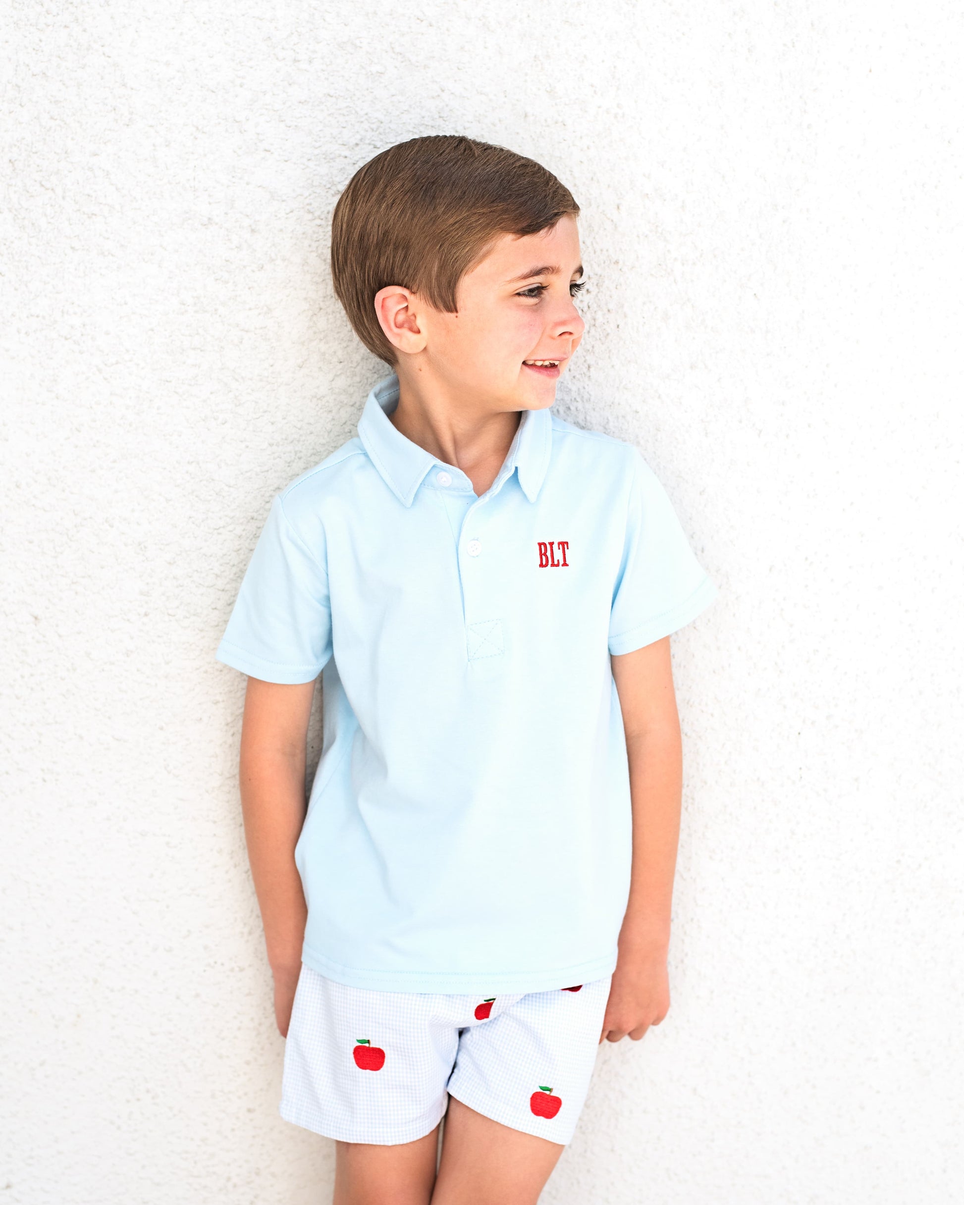 Young boy wearing blue poo shirt and blue gingham shorts with apples embroidered.