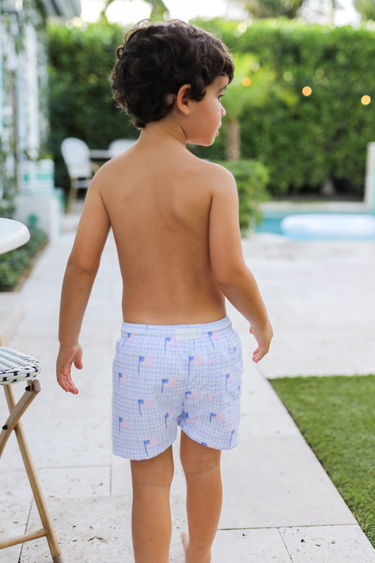 Young boy wearing checkered swim shorts walking outdoors near a pool.