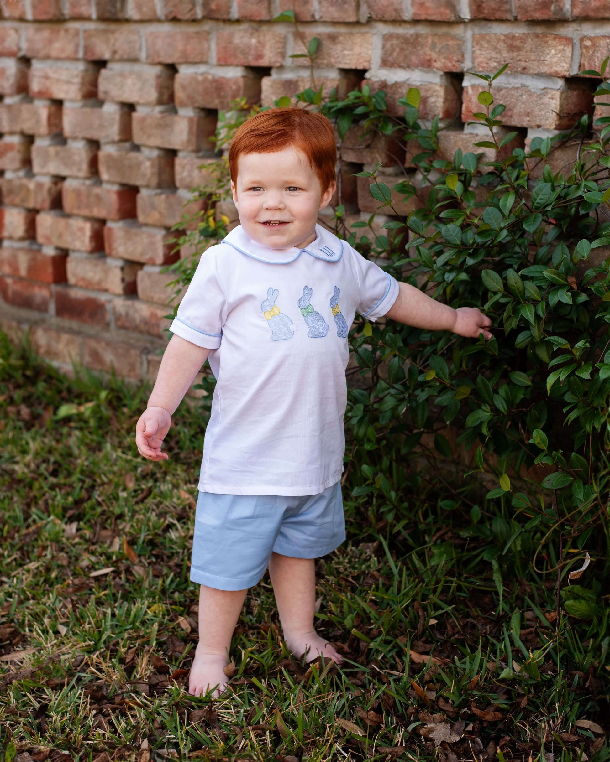 Young boy with red hair standing outdoors near a brick wall and greenery