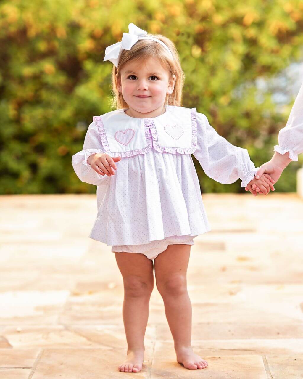 Young child in a white dot diaper set with ruffles standing on a wooden floor outdoors.