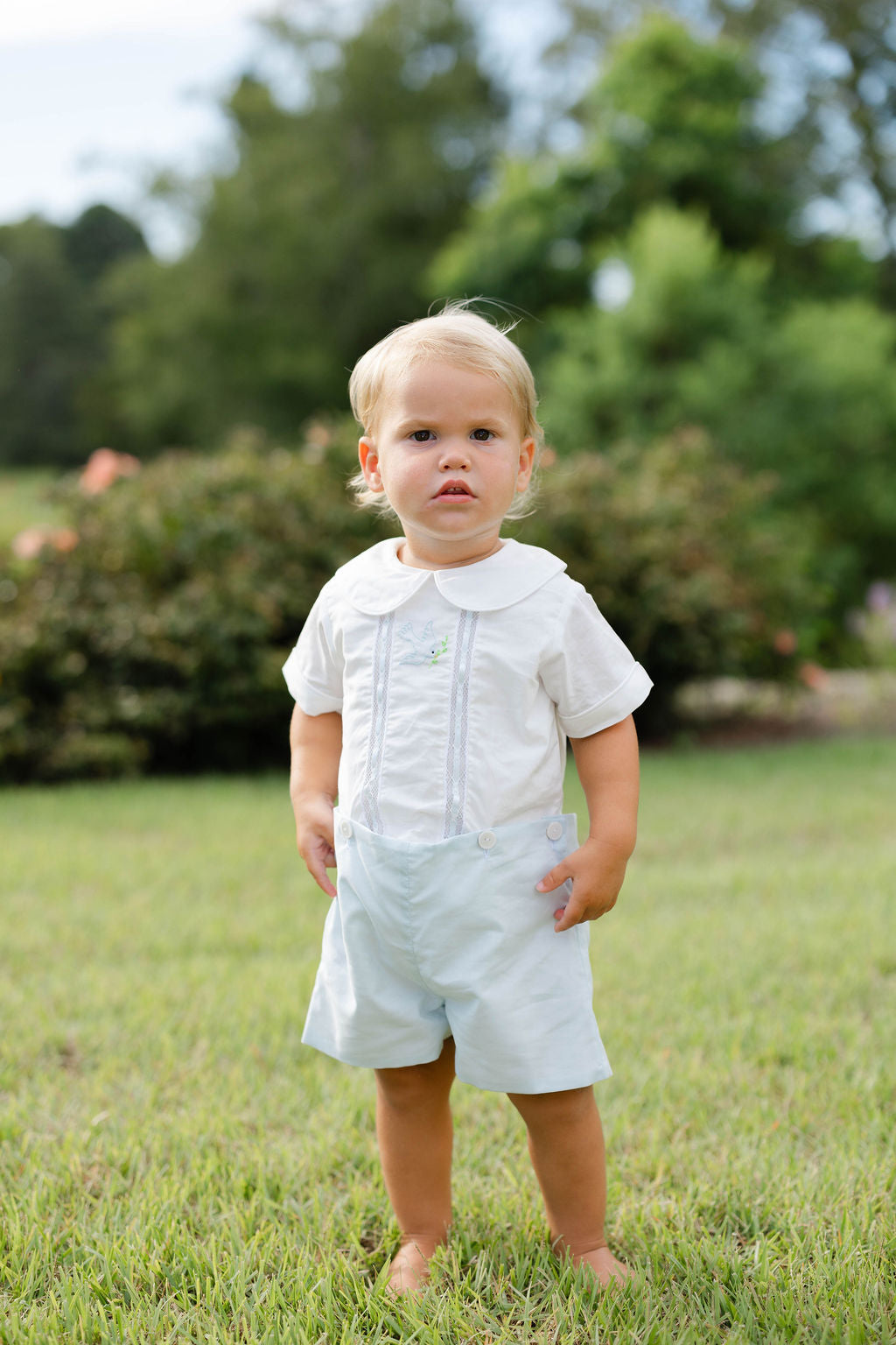 Young child in a white shirt and light blue shorts standing on grass with trees in the background