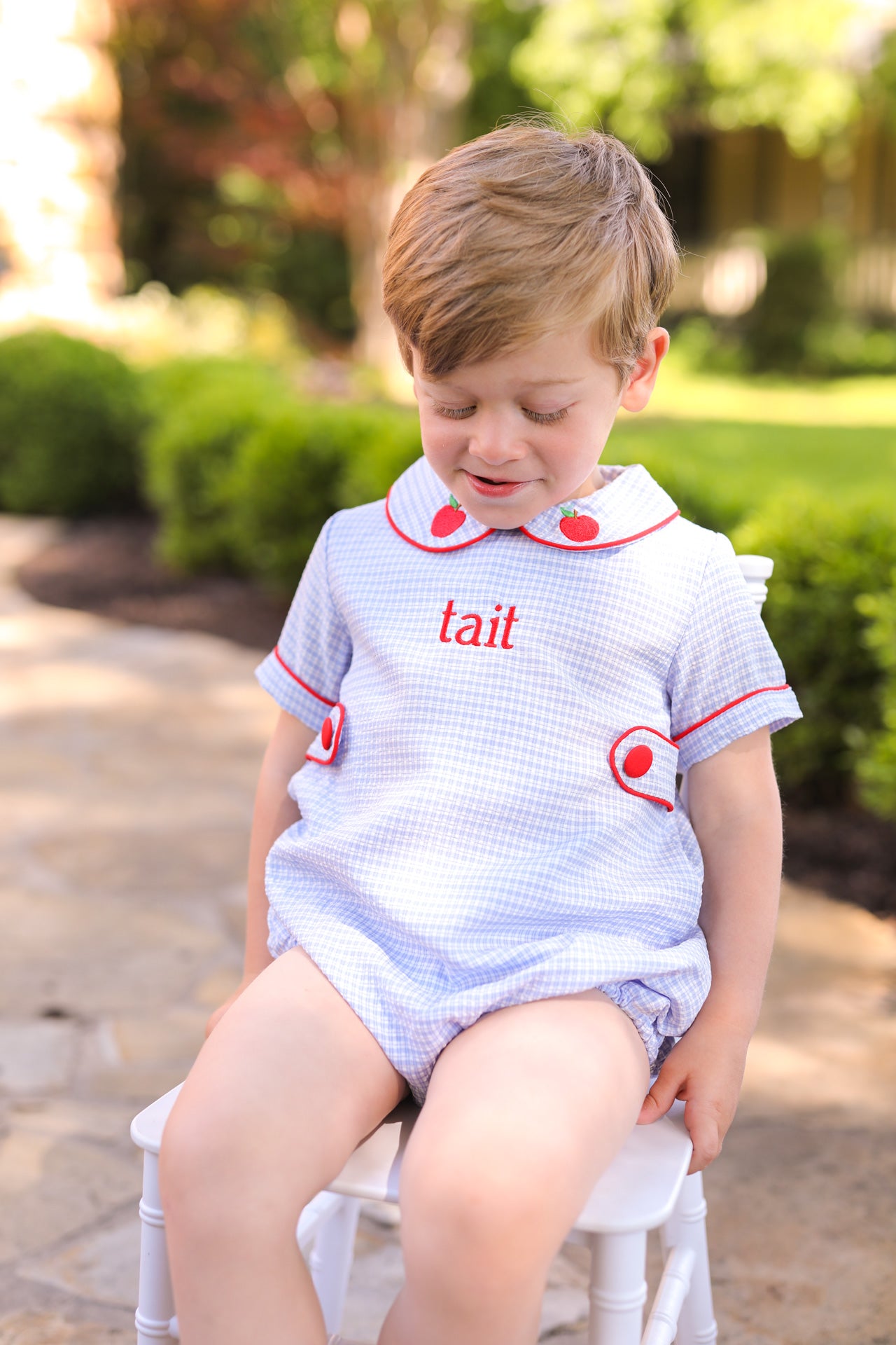 Young child sitting on a chair wearing a blue outfit with apples on the collar.