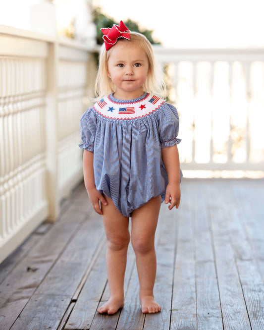 Young child wearing a blue checkered romper with red and white accents on a wooden deck.