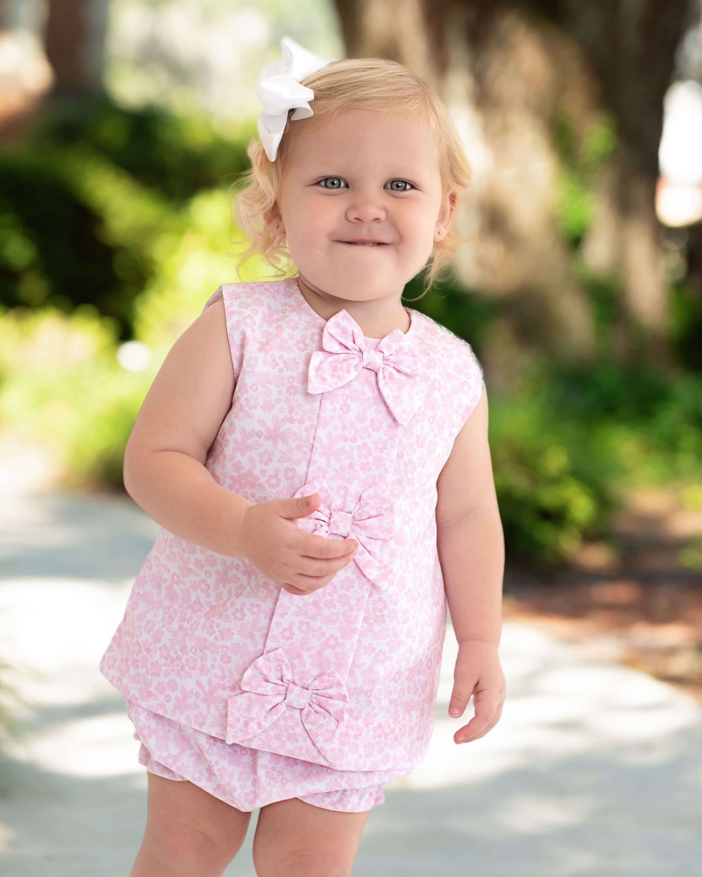 Young child wearing a pink floral outfit with a bow, standing outdoors.
