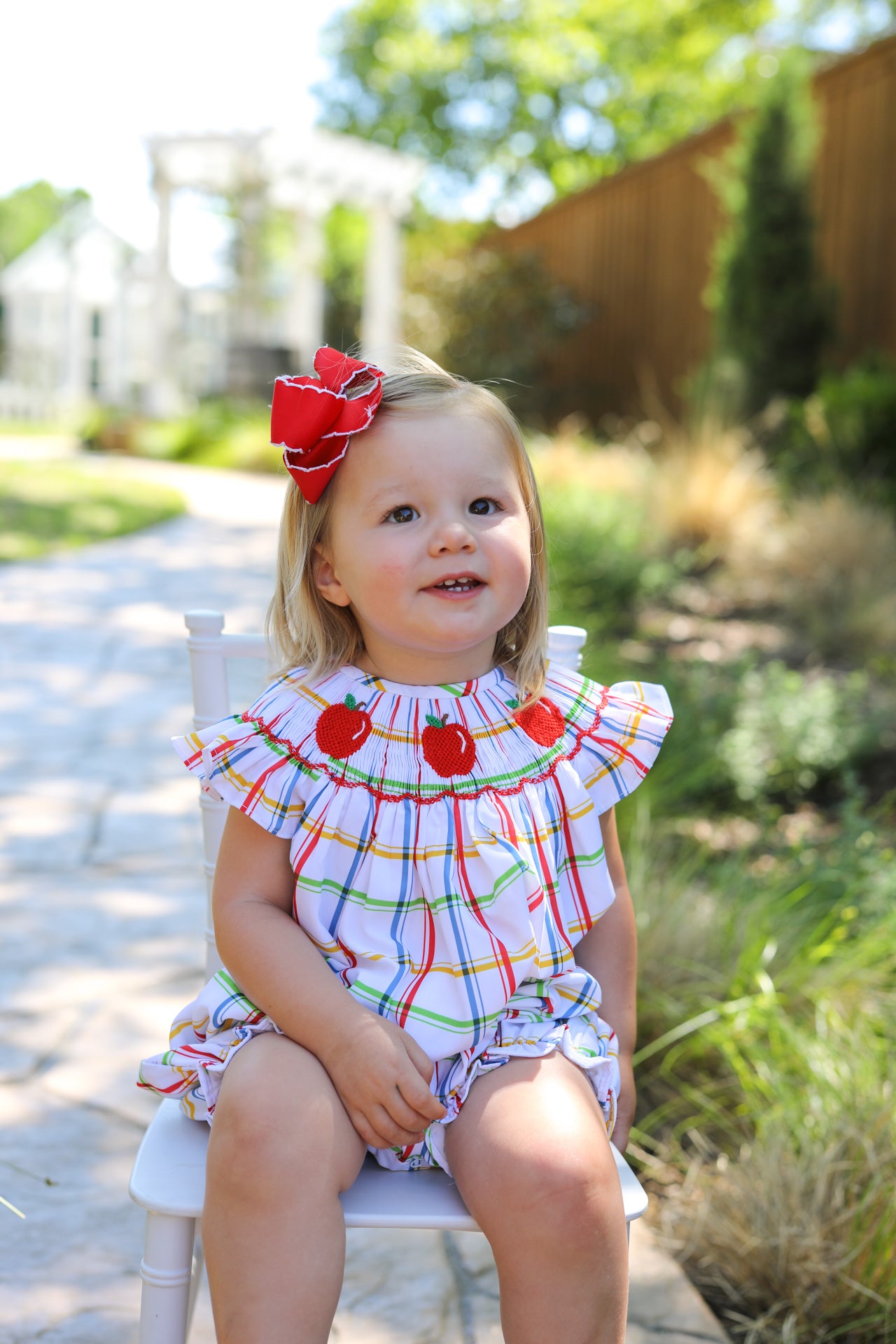 Young child wearing plaid outfit with smocked apples sitting on a chair.