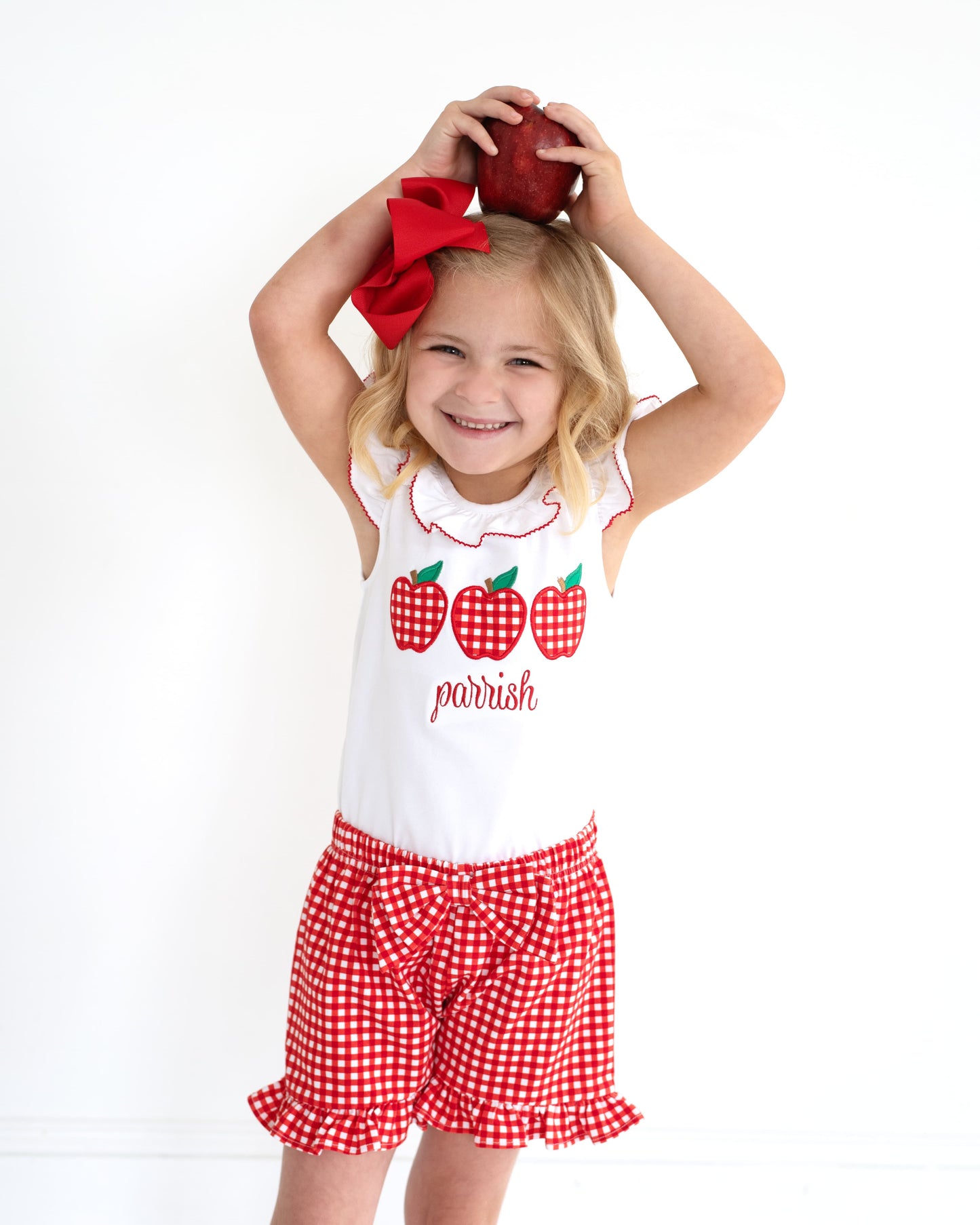 Young girl holding an apple over her head wearing red gingham shorts and an a shirt with apples.
