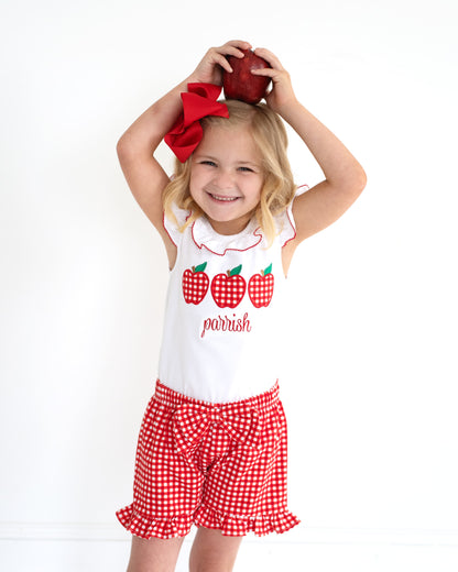 Young girl holding an apple over her head wearing red gingham shorts and an a shirt with apples.