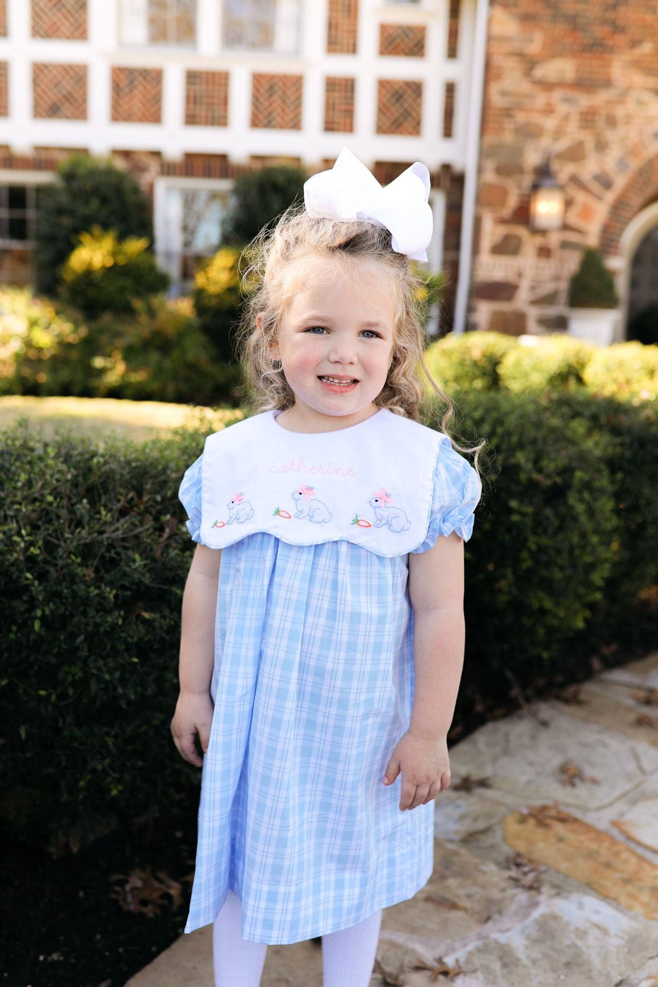 Young girl in a blue checkered dress with a white bow standing outdoors.