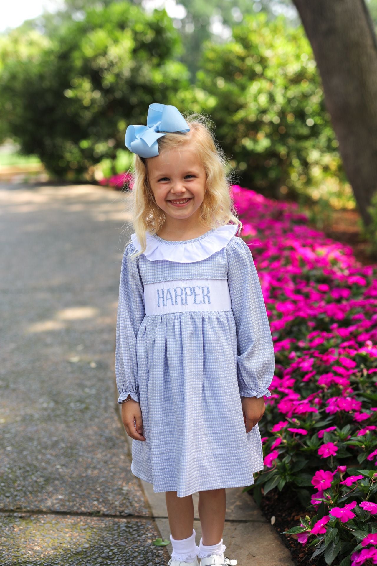 Young girl in a blue dress with a bow, standing outdoors near pink flowers.