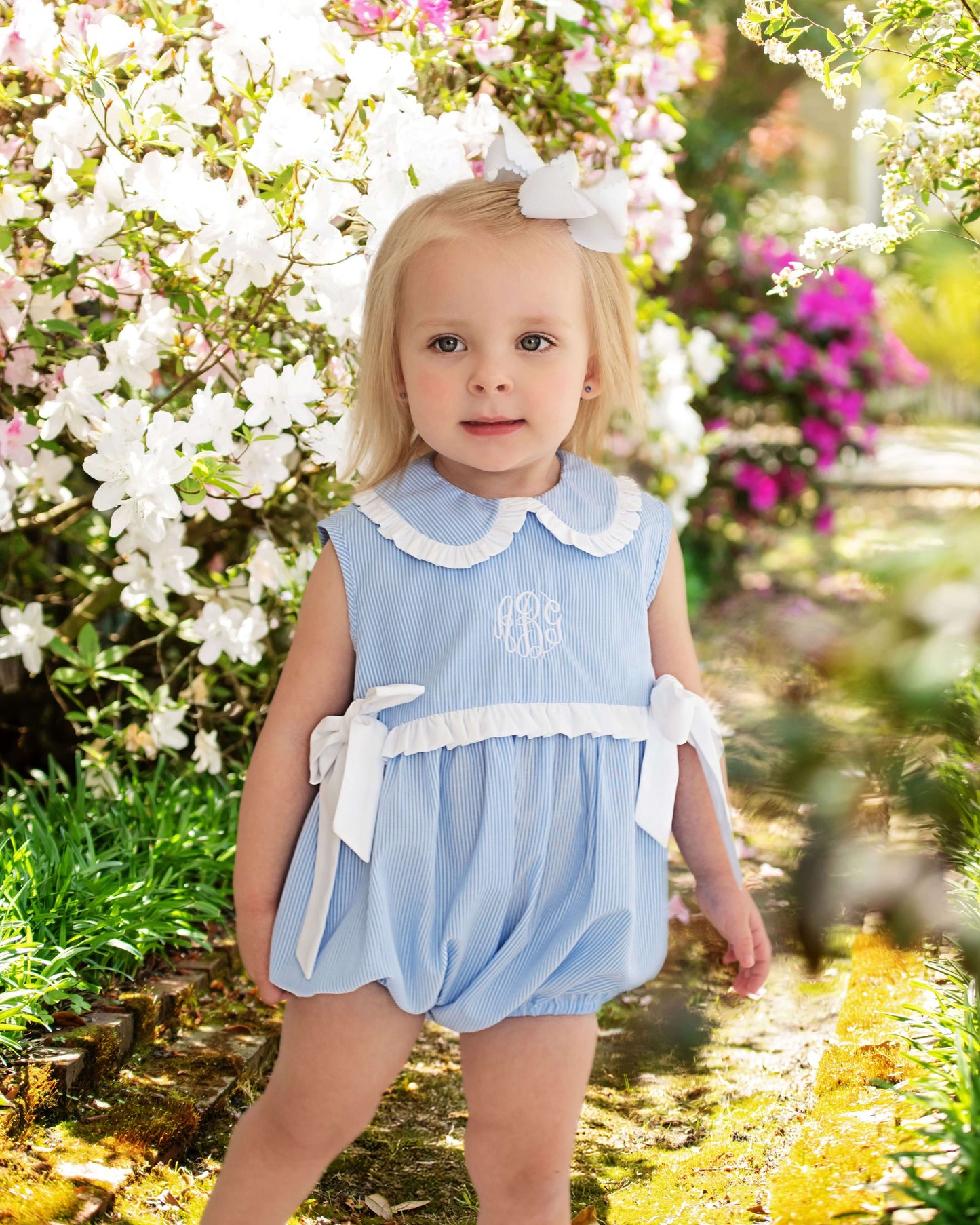 Young girl in a blue dress with white trim standing in a garden with flowers and a pond.