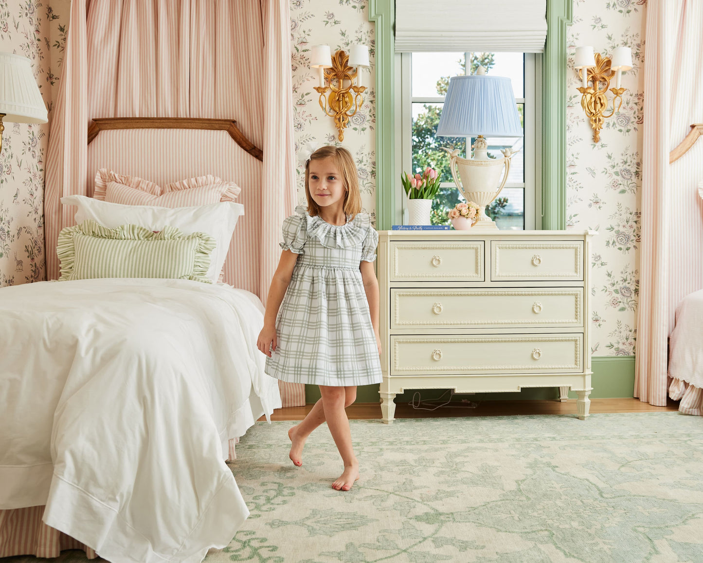 Young girl in a checkered dress standing in a decorated bedroom with a bed and dresser.