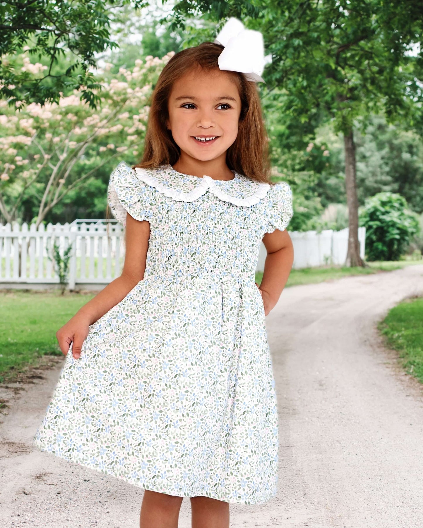 Young girl in a floral dress standing on a path with trees and a white fence in the background
