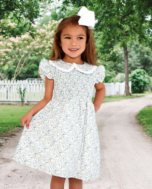 Young girl in a floral dress standing on a path with trees and a white fence in the background