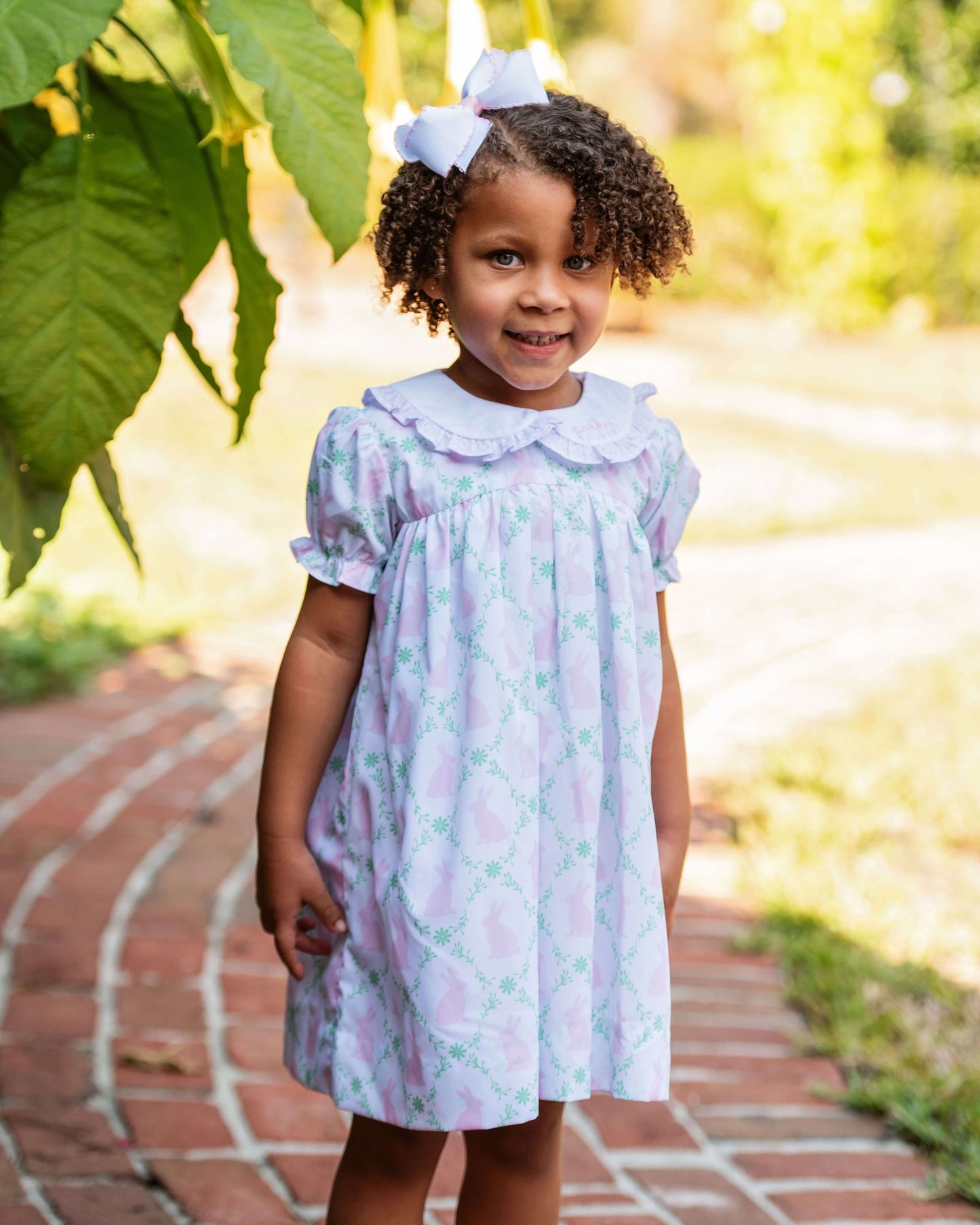 Young girl in a floral dress standing outdoors on a brick path with greenery.