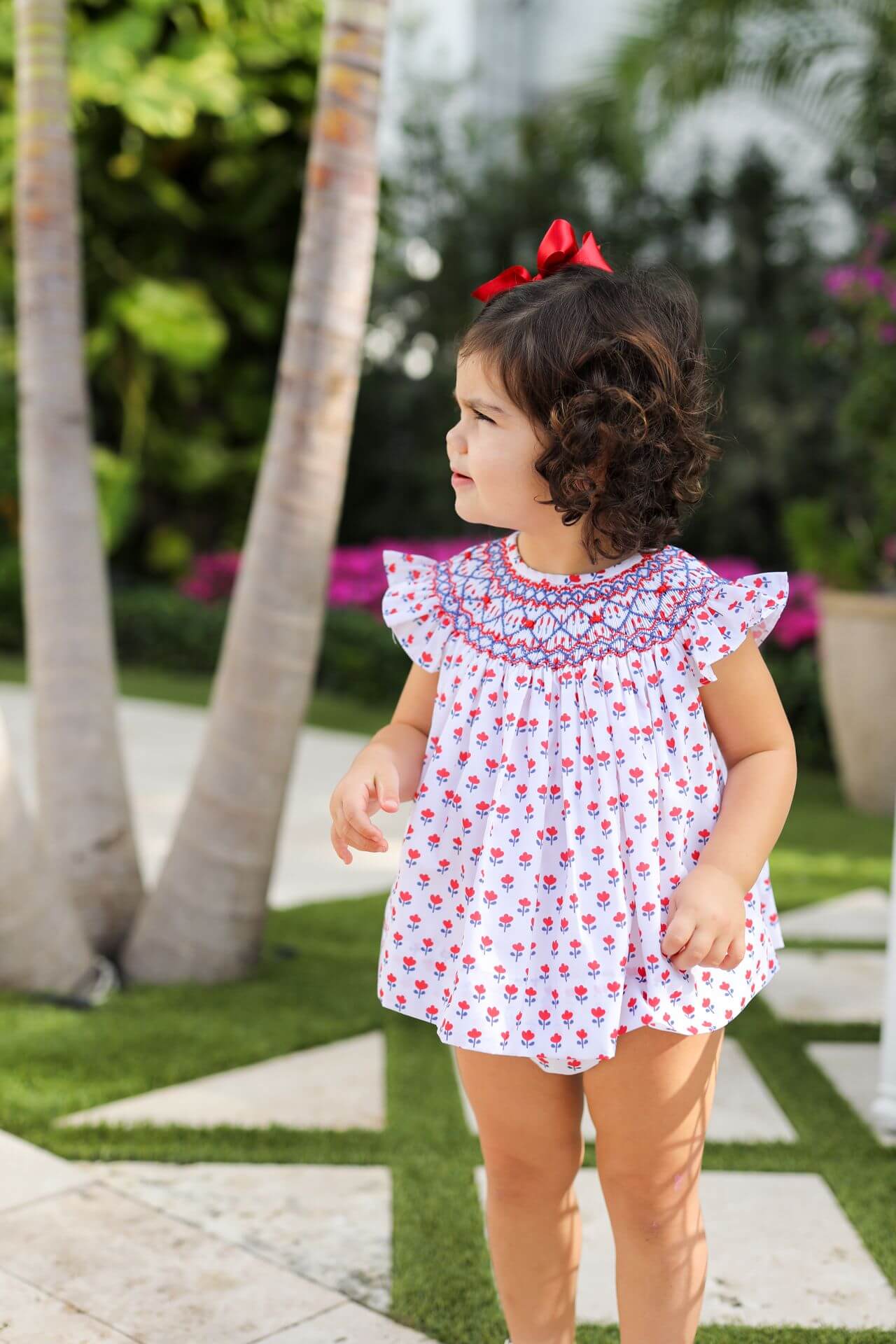 Young girl in a floral dress standing outdoors with palm trees and greenery in the background