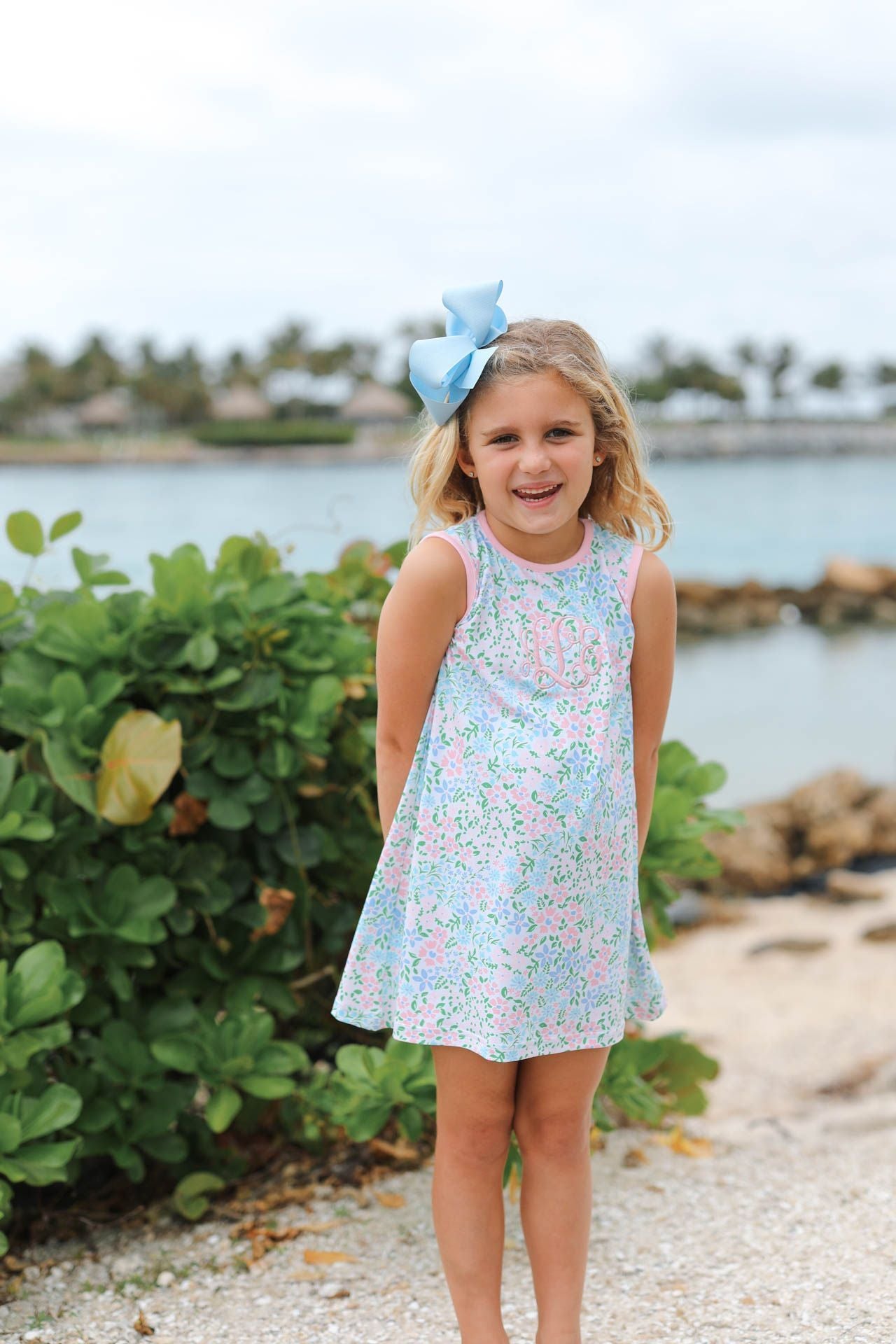 Young girl in a floral dress with a blue bow standing on a beach.