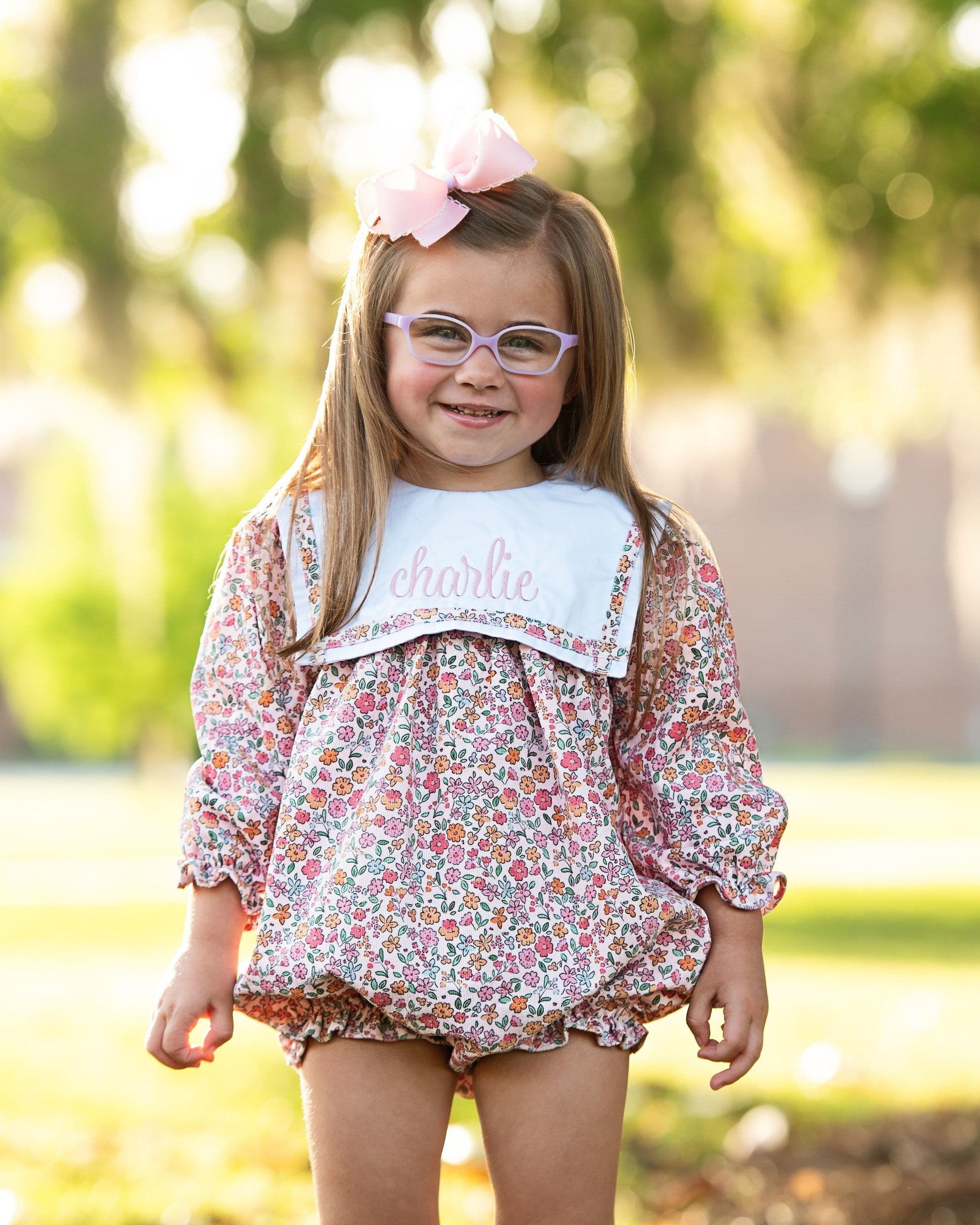 Young girl in a floral dress with a bow and glasses, standing outdoors.