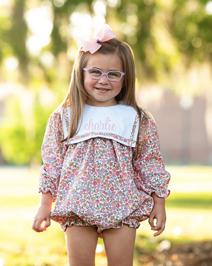 Young girl in a floral dress with a bow and glasses, standing outdoors.