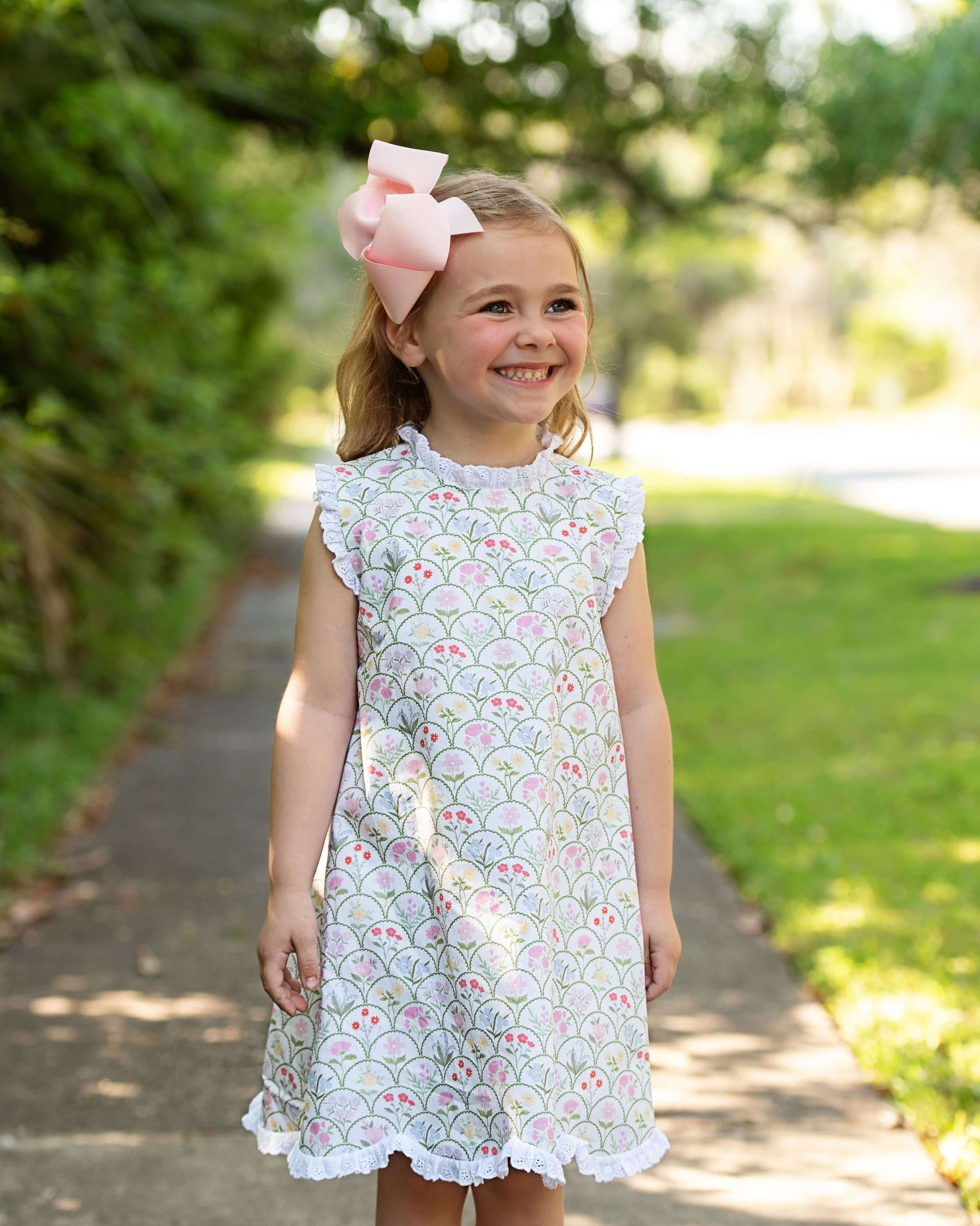 Young girl in a floral dress with a pink bow standing outdoors on a sunny day.