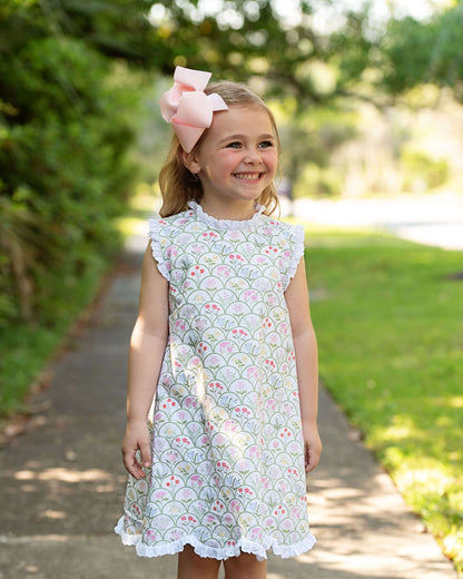 Young girl in a floral dress with a pink bow standing outdoors on a sunny day.