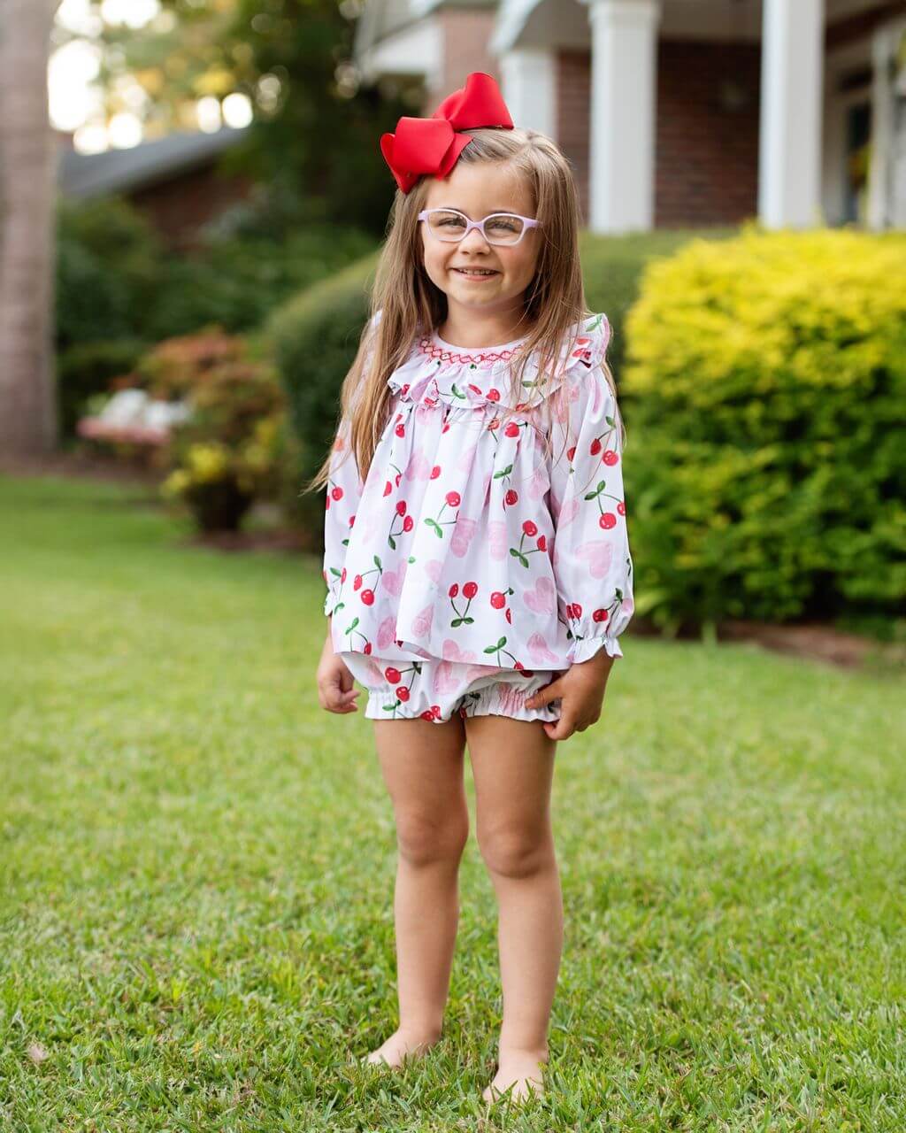 Young girl in a floral dress with cherries and a red bow in her hair, standing on grass.