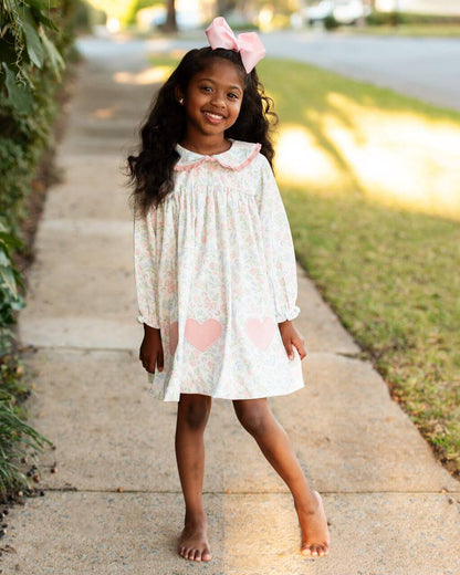 Young girl in a floral dress with pink hearts standing on a sidewalk.