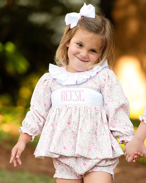 Young girl in a floral outfit with a bow, standing outdoors.
