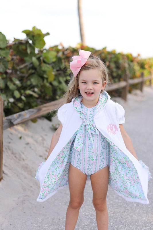 Young girl in a floral swimsuit with a white cover-up and pink bow, standing on a beach.