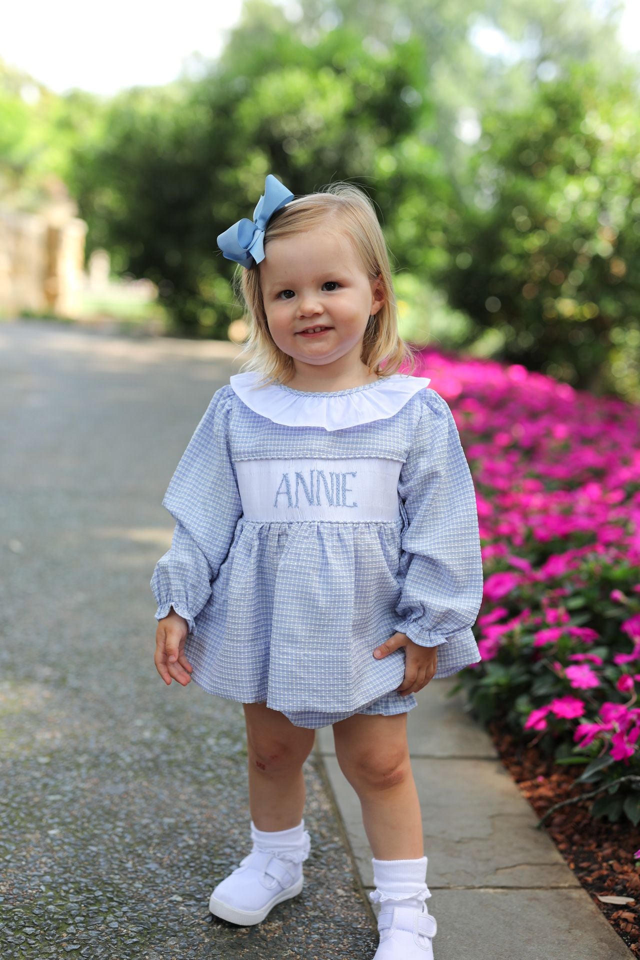 Young girl in a gray dress with 'ANNIE' embroidered on it, standing outdoors with greenery and flowers in the background.