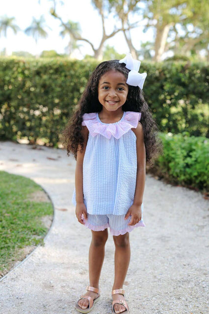 Young girl in a light blue and white dress with a large bow standing on a path outdoors.