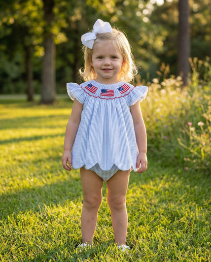 Young girl in a light blue dress with American flag designs standing in a grassy field.